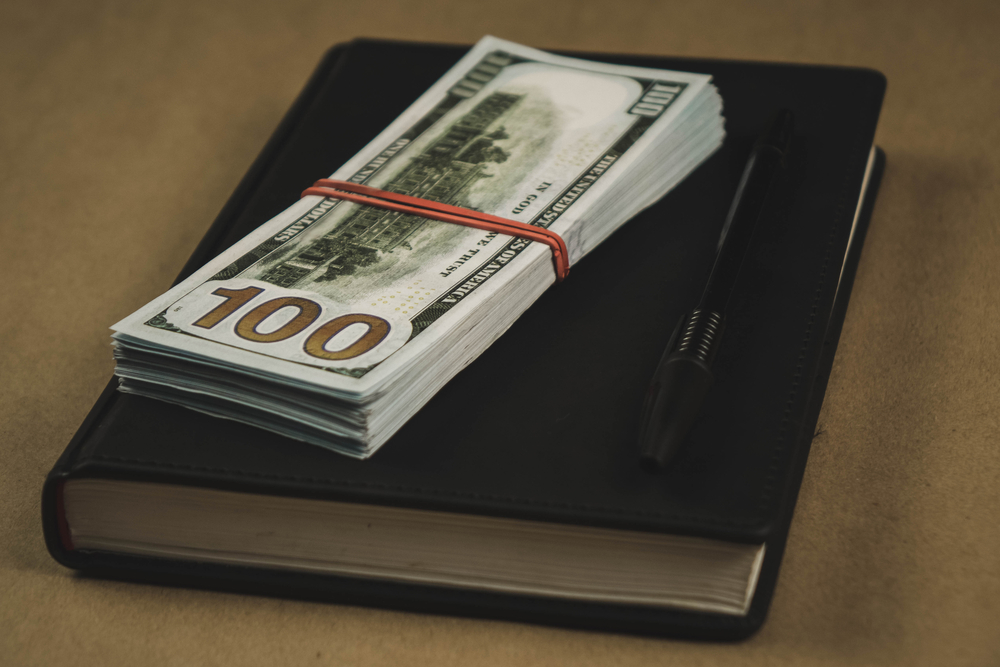 A black notebook , a pen and dollar cash banknotes on wooden background - concept of financial management or planning, make money from freelance writing